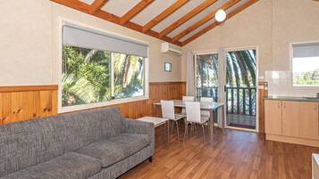 A room with a wooden ceiling, a grey sofa, a dining table with chairs, and a kitchen area with wooden cabinets.