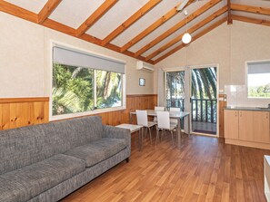 A room with a wooden ceiling, a grey sofa, a dining table with chairs, and a kitchen area with wooden cabinets.