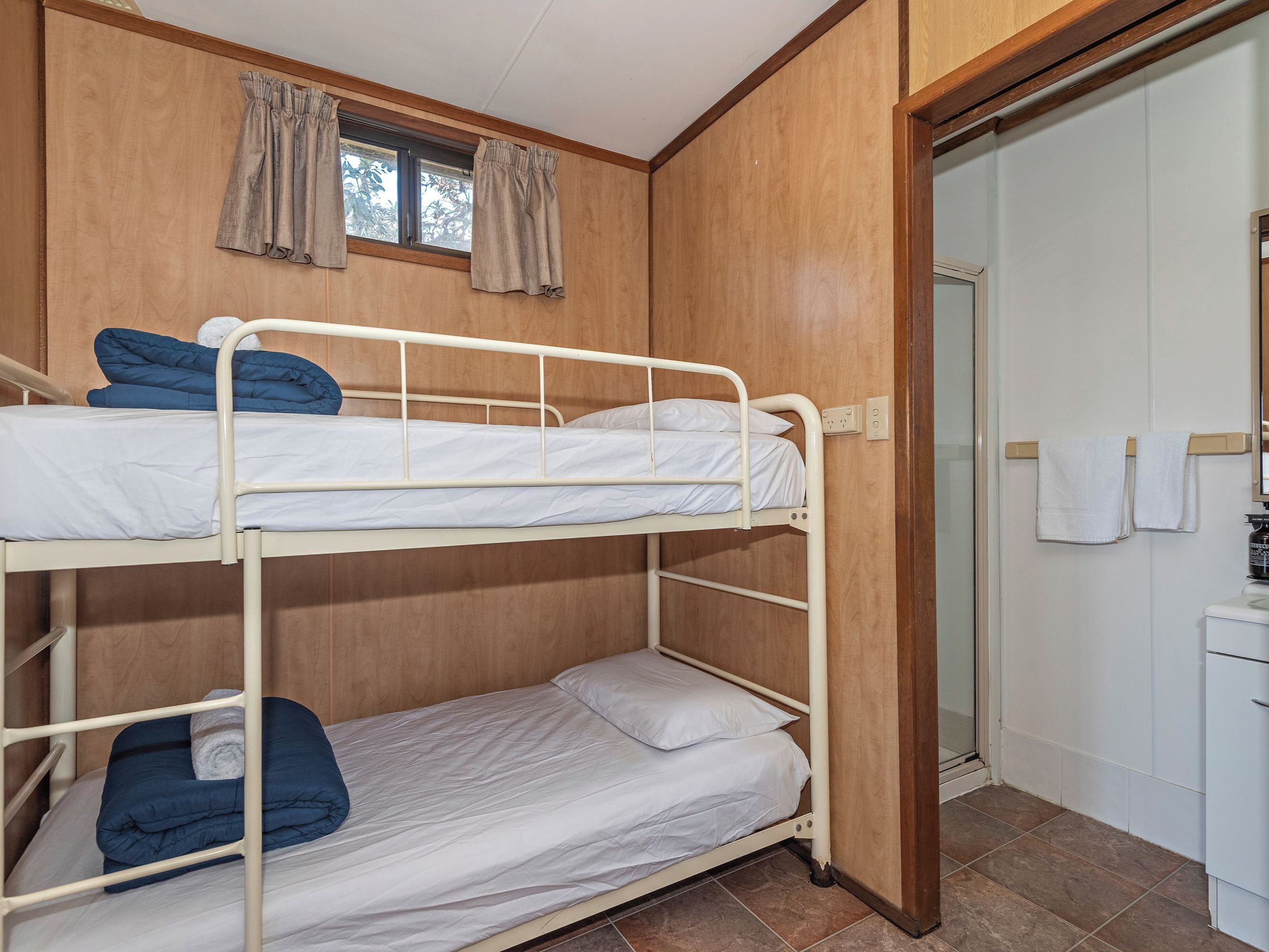 A bunk bed room with a wooden wall, a window with curtains, and a door leading to a bathroom.