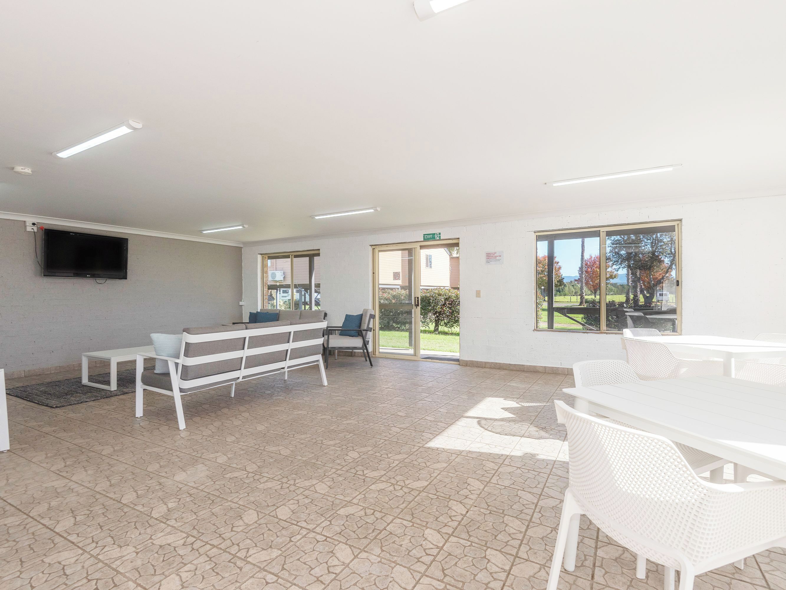 Un espacio interior amplio con un televisor de pantalla plana montado en la pared, una zona de estar con muebles blancos y un ventanal que ofrece vistas al exterior.