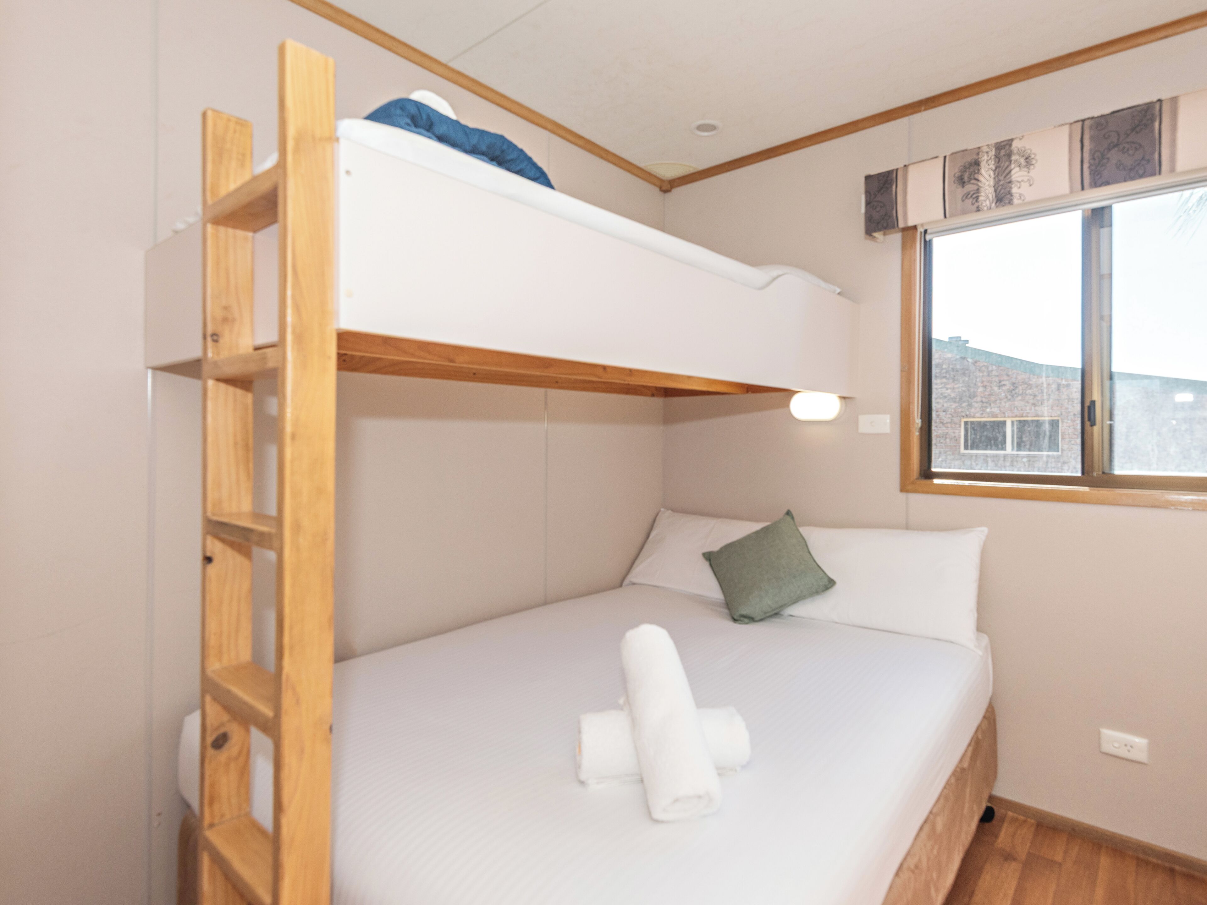 Una litera con escalera de madera, una cama individual con almohada y una ventana con vista a edificios.