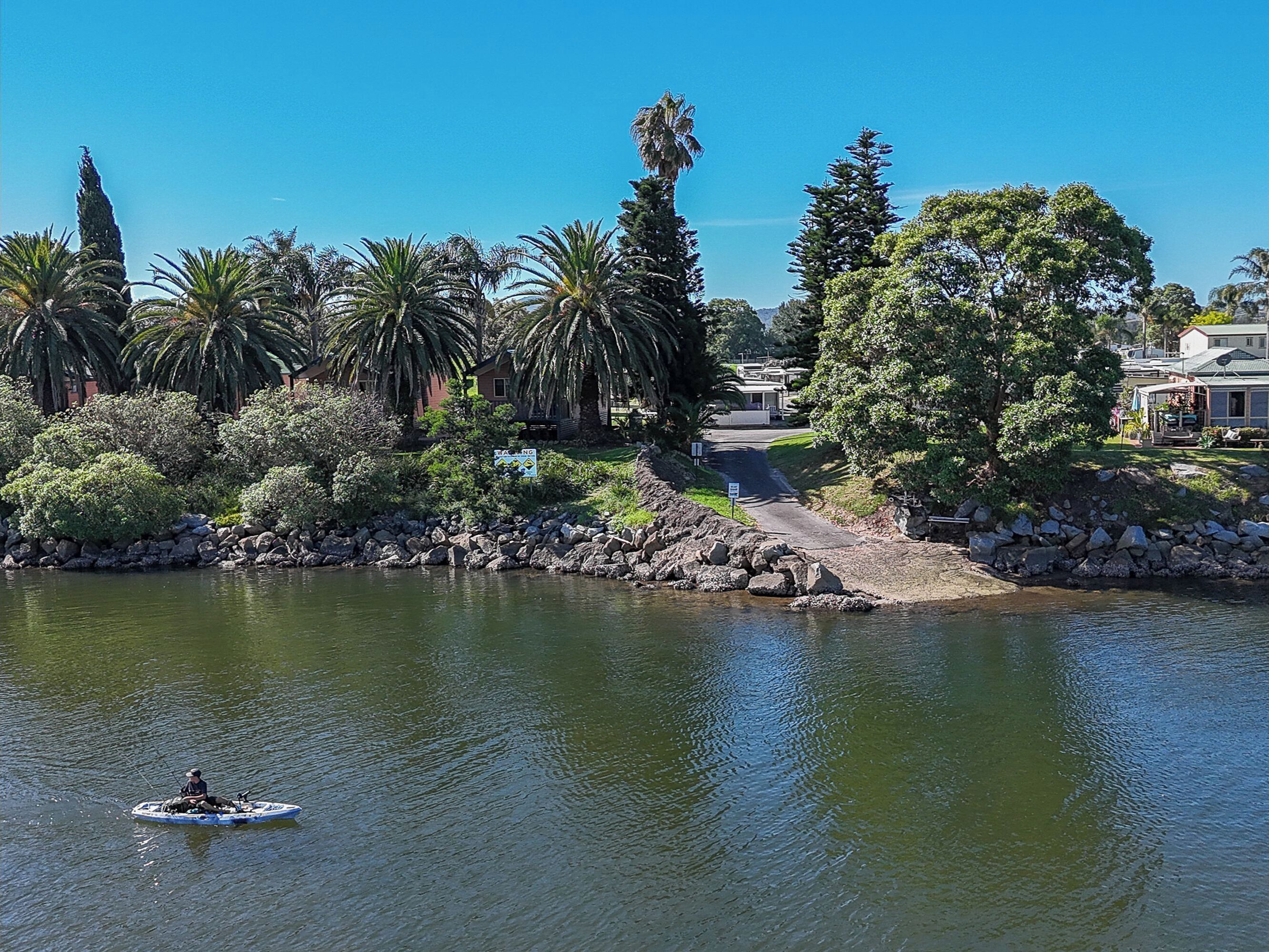 Une rivière paisible où une personne pêche dans un petit bateau, entourée d’une végétation luxuriante et sous un ciel bleu clair.