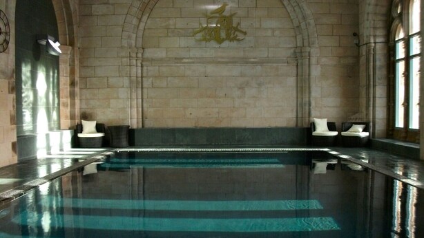Ein großer, überdachter Pool mit Steinwand und Rundbogenzudecke, zwei Liegestühle und ein dekoratives Wappen über dem Pool.