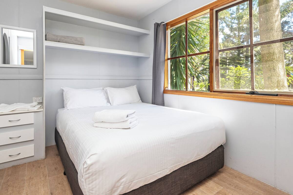 A bedroom with a bed, built-in shelving, a window with a view of trees, and a wooden floor.