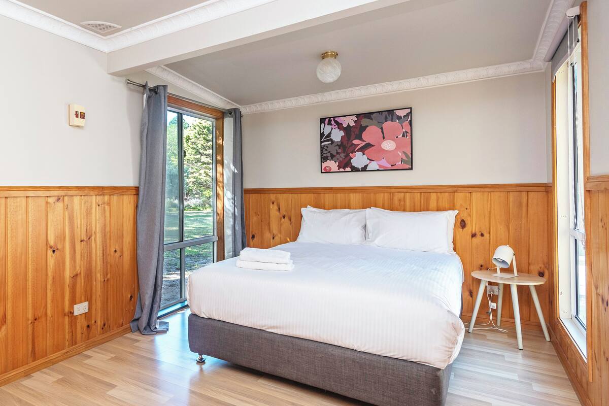 A bedroom with a bed, wooden paneling, a window with curtains, a small table with a lamp, and a floral painting on the wall.