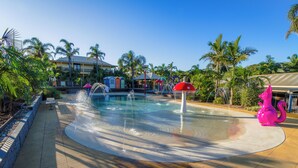 A pool area with a water feature, palm trees, and a building with colorful beach huts in the background. - Wairo Beach Holiday Park (Lake Tabourie)