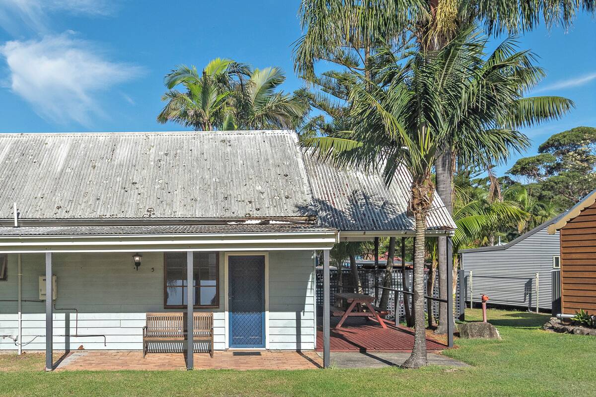 A single-story building with a corrugated metal roof, a wooden bench outside, and a palm tree nearby.