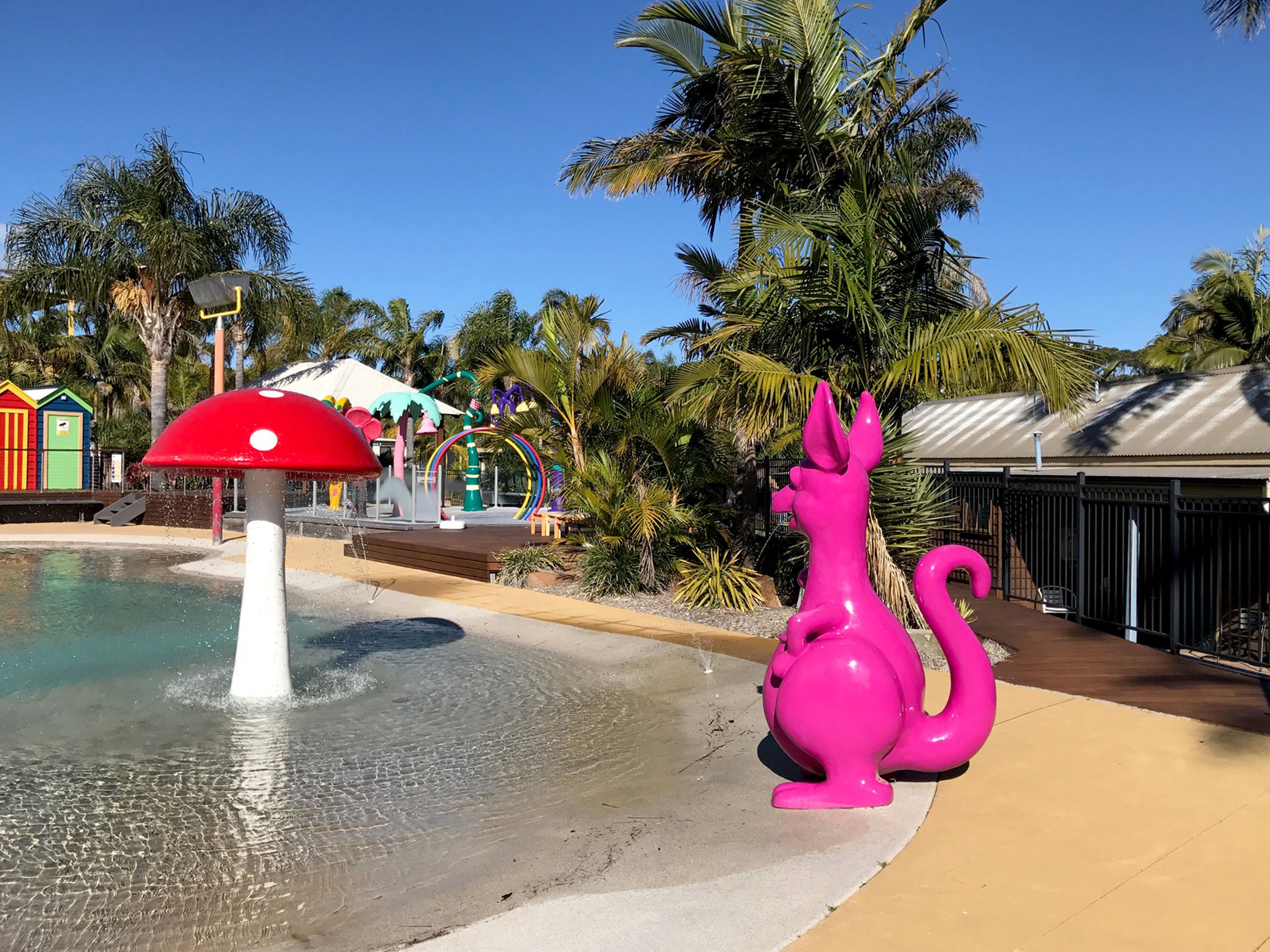 A water play area with a red mushroom-shaped fountain, a pink sculpture, and palm trees in the background.