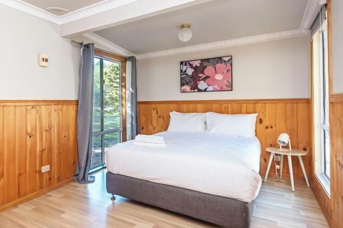 Ein Schlafzimmer mit einer Wandpaneele aus Holz, einem Bett mit weißen Leinen, einem kleinen Nachttisch und einem Fenster mit Blick.