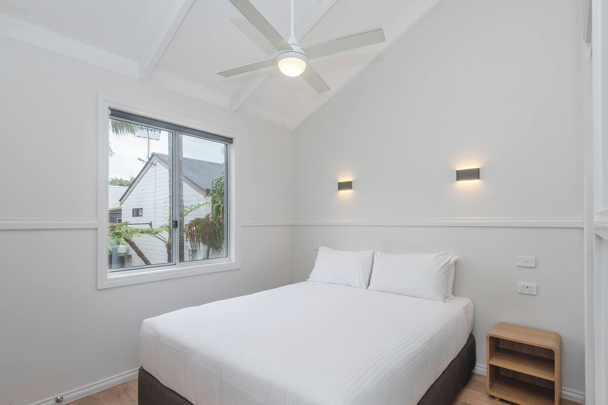 A bedroom with a bed, a ceiling fan, a window, and a nightstand.