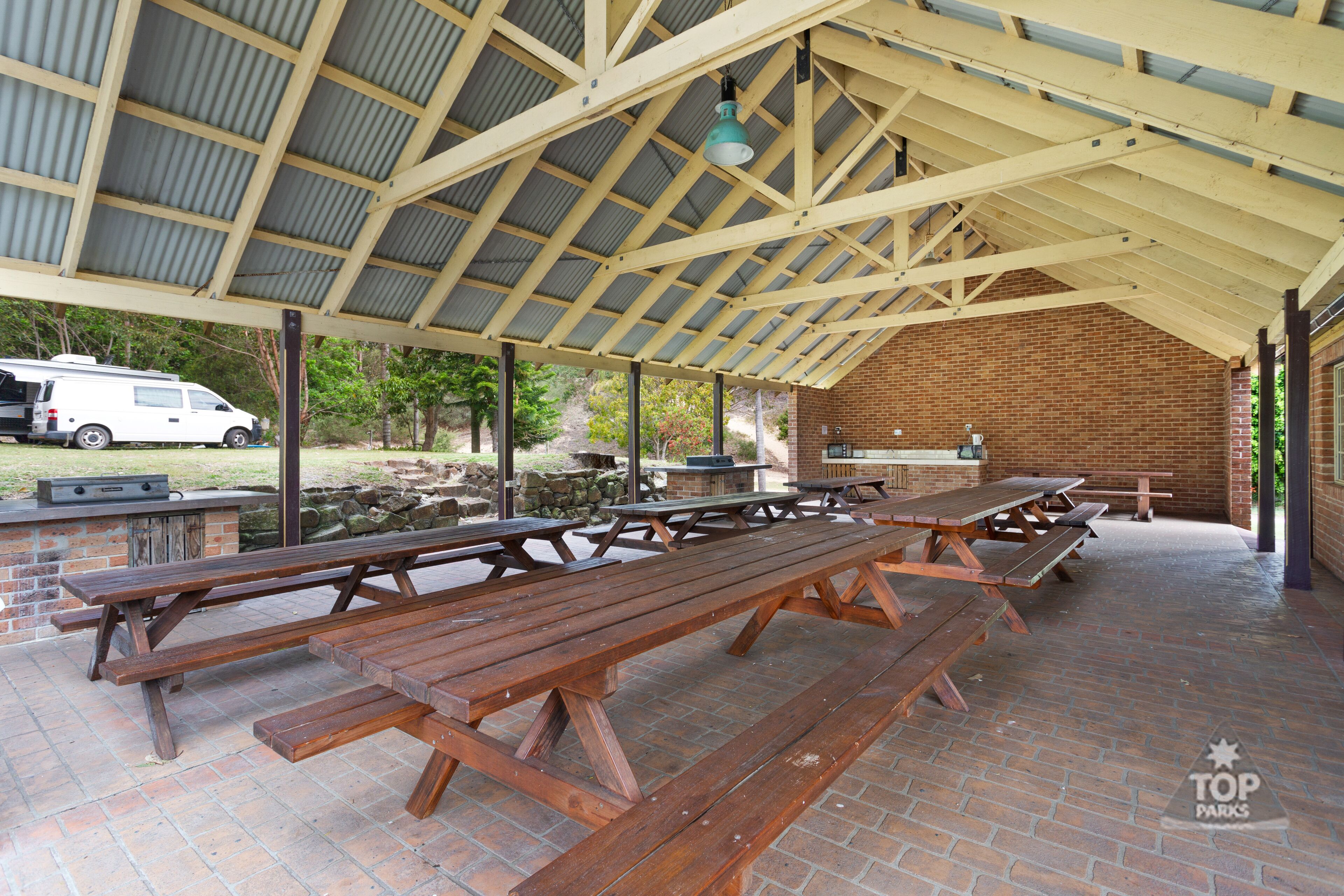 A covered picnic area with multiple wooden picnic tables and a barbecue grill.