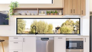 A modern kitchen with a dishwasher, microwave, and a large window overlooking a garden. - Wairo Beach Holiday Park (Lake Tabourie)