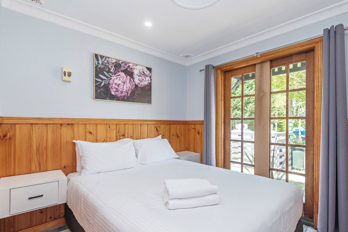 A hotel room with a wooden headboard, a large bed with white linens, a nightstand, a wall-mounted light, and a window with curtains.