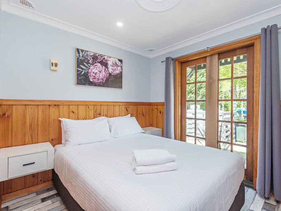 A hotel room with a wooden headboard, a large bed with white linens, a nightstand, a wall-mounted light, and a window with curtains.