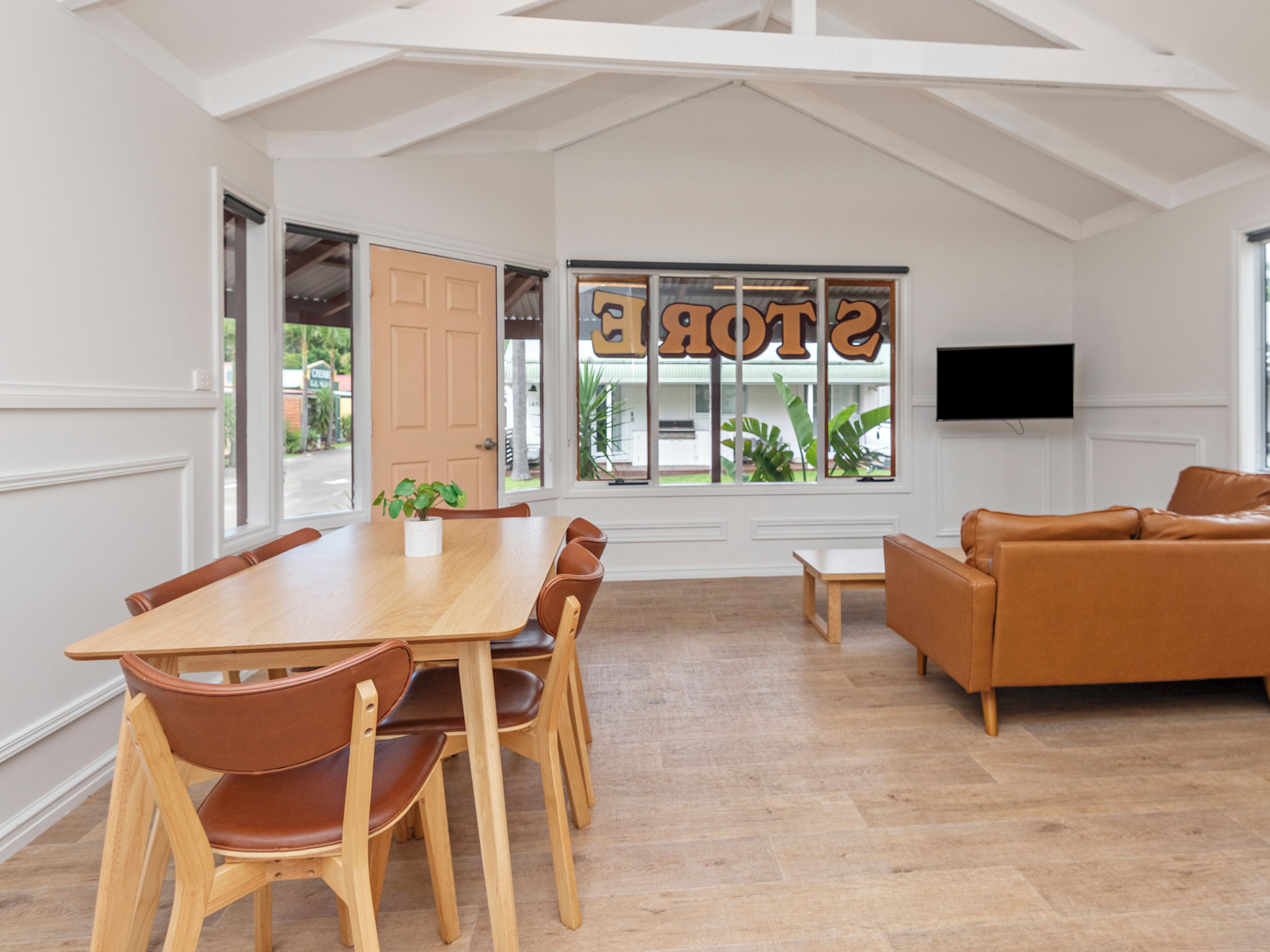 A dining area with a wooden table and chairs, a sofa, a television, and a window with a view of greenery.