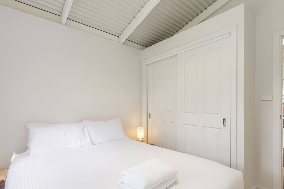 A bedroom with a bed, pillows, a folded towel, a nightstand, and a closed door.