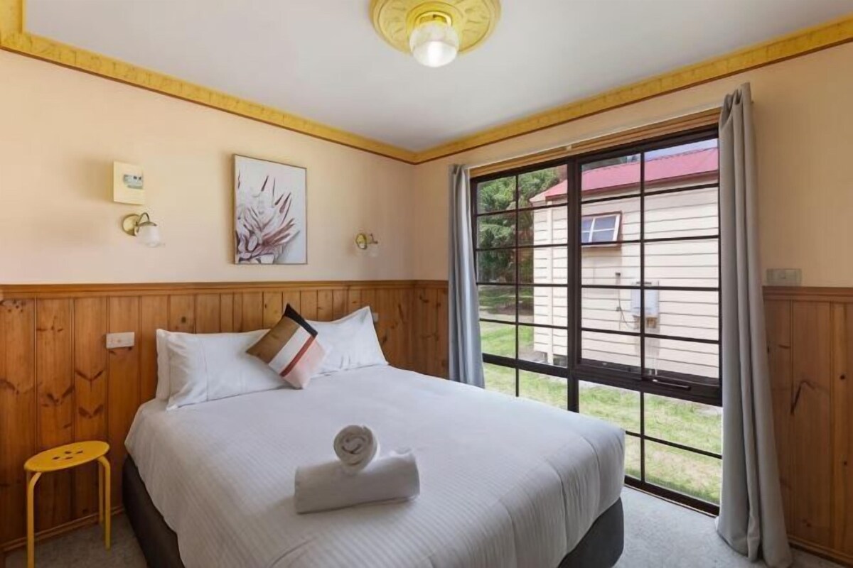 Ein Schlafzimmer mit einem großen Bett, einem kleinen Hocker, einem Fenster mit Vorhängen und einer Wandleuchte.
