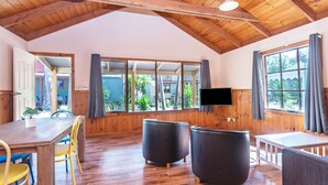 A room with wooden beams, a TV mounted on the wall, and a large window with curtains. - Wairo Beach Holiday Park (Lake Tabourie)