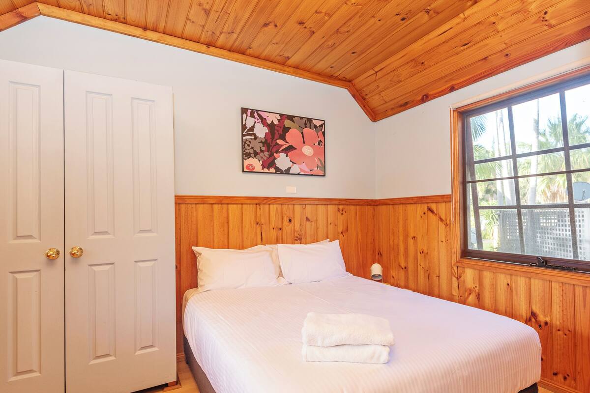 A bedroom with a wooden ceiling, a bed with white bedding, a window with a view of palm trees, and a painting on the wall.