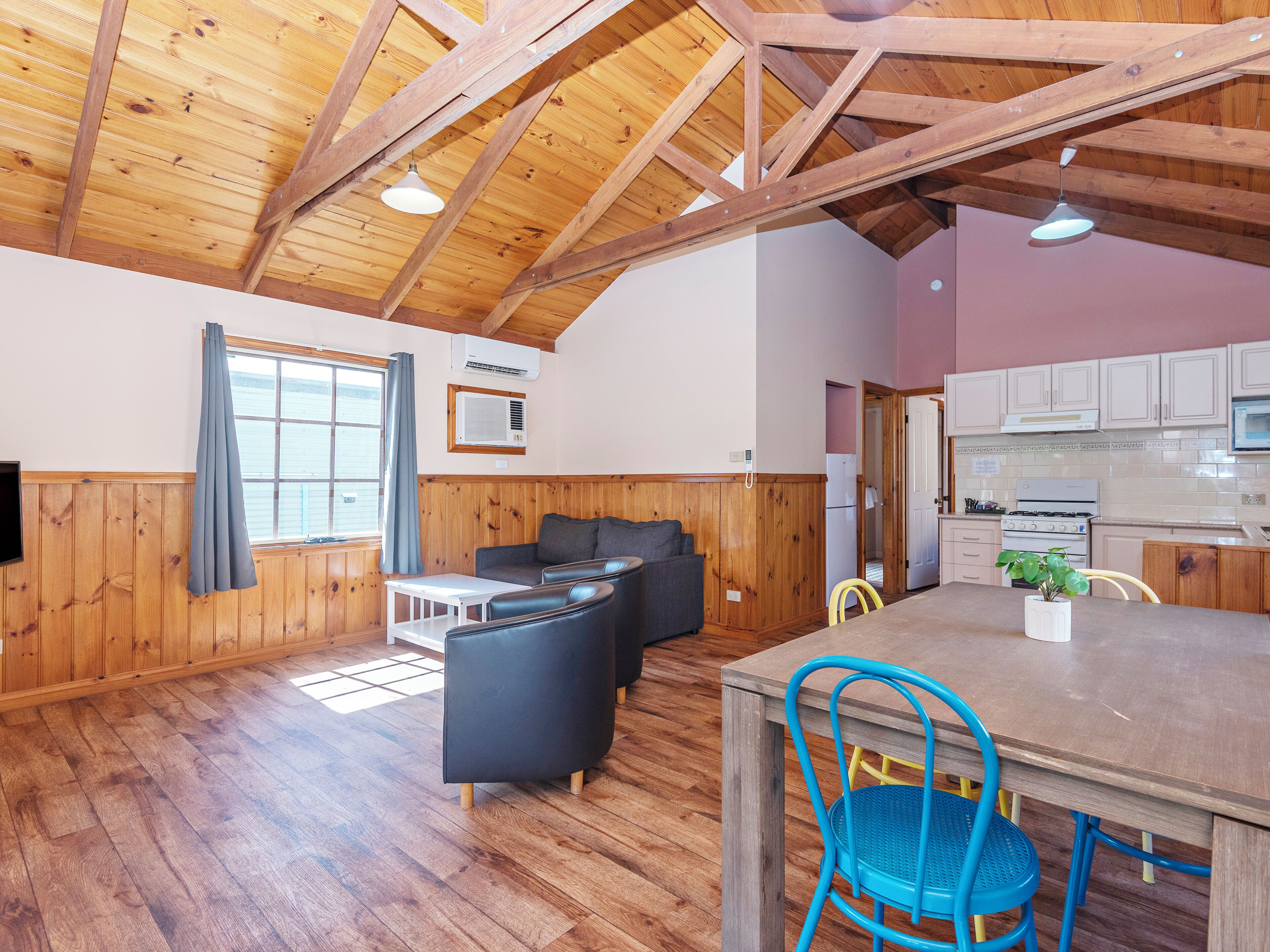 A cozy cabin interior with wooden walls, a dining area, a kitchen, and a living room with a couch and chairs.