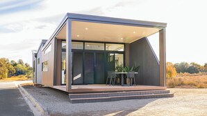 A modern house with a large glass door, a wooden deck, and a table with chairs outside. - Wairo Beach Holiday Park (Lake Tabourie)