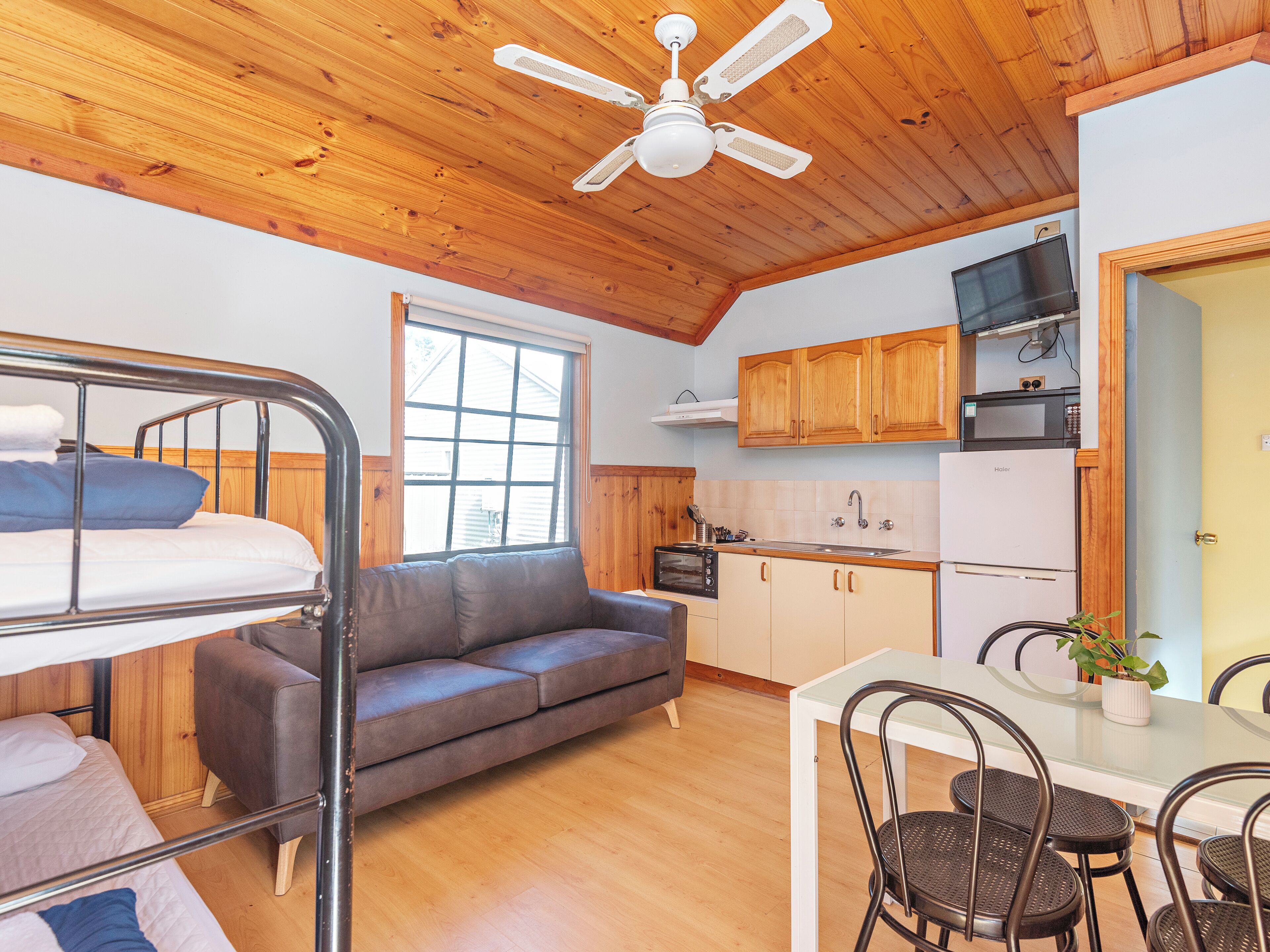 A compact room with bunk beds, a sofa, a dining table with chairs, a kitchen area with appliances, and a ceiling fan.
