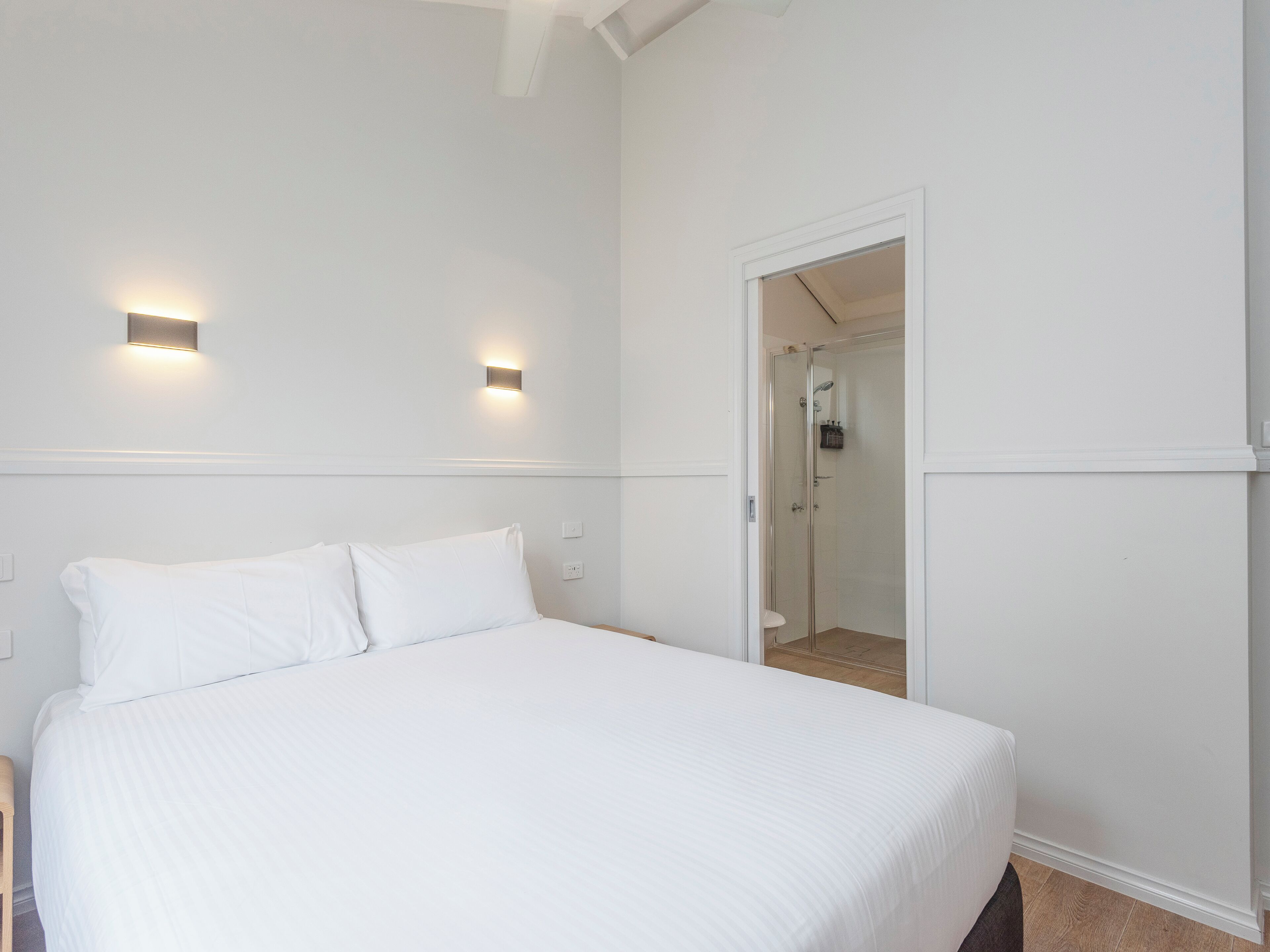 Een hotelkamer met een bed, twee wandlampen en een douche met glazen wanden.
