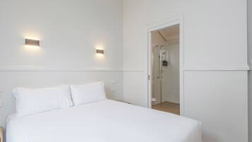 Quarto de hotel com uma cama, duas luminárias fixadas na parede e um chuveiro com divisória de vidro.