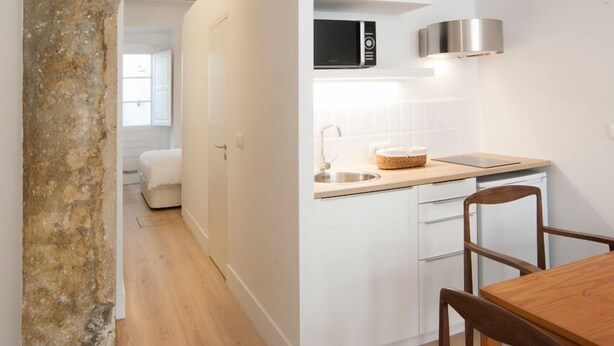 Une cuisine compacte avec des meubles blancs, une table à manger en bois et un arc en pierre menant à une chambre.
