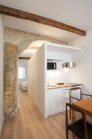 Cocina compacta con armarios blancos, una mesa de comedor de madera y un arco de piedra que conduce a un dormitorio.