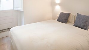 Une chambre avec un grand lit, une poutre en bois au plafond, une lampe fixée au mur et une fenêtre dont les volets sont ouverts.