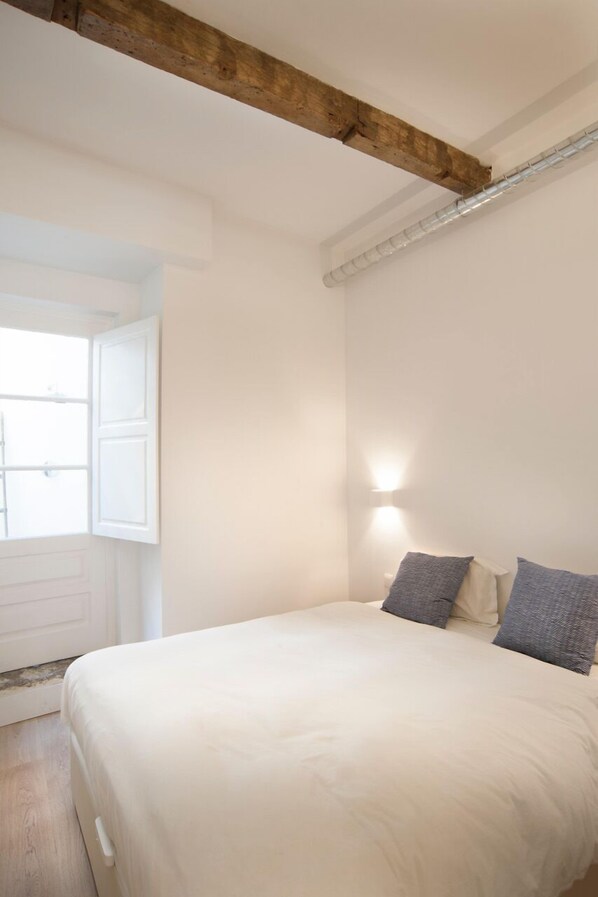 Un dormitorio con una cama grande, una viga de techo de madera, una lámpara de pared y una ventana con contraventanas abiertas.
