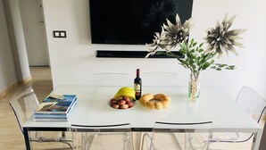 Et moderne spiseområde med et hvidt bord, gennemsigtige stole, et stort fladskærms-tv monteret over, og dekorative planter.