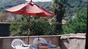 A table set outdoors with a red umbrella, white chairs, and a view of a mountainous landscape. - Beautiful village home in the foothills of the Pyrenees (Finestret)