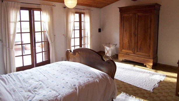 A bedroom with a wooden ceiling, a large bed with a floral bedspread, a wooden wardrobe, and a window with curtains. - Beautiful village home in the foothills of the Pyrenees (Finestret)