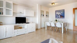 Ein modernes Wohnzimmer mit Fernseher, weißen Schränken, einem Essbereich mit Tisch und Stühlen sowie einem Bild an der Wand.