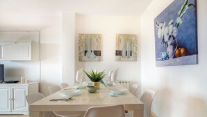 Ein Essbereich mit einem weißen Tisch, gedeckt für vier Personen, umgeben von weißen Stühlen. An der Wand hängen zwei Gemälde, eines zeigt Blumen, das andere abstrakte Formen.