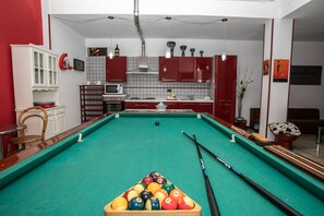 A pool table in a room with a kitchen area, including cabinets and a microwave.