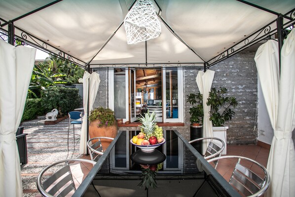 A covered outdoor dining area with a table set for two, surrounded by metal chairs, and a hanging light fixture above.