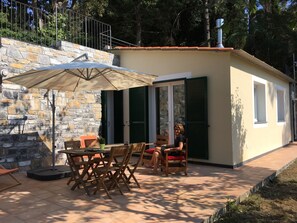 A stone wall with a wooden table and chairs, a large umbrella, and a small building with green shutters.