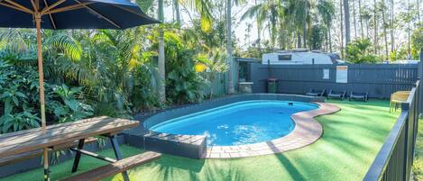 Una piscina con cubierta de madera y una sombrilla, rodeada de exuberante vegetación y una zona delimitada por una valla.