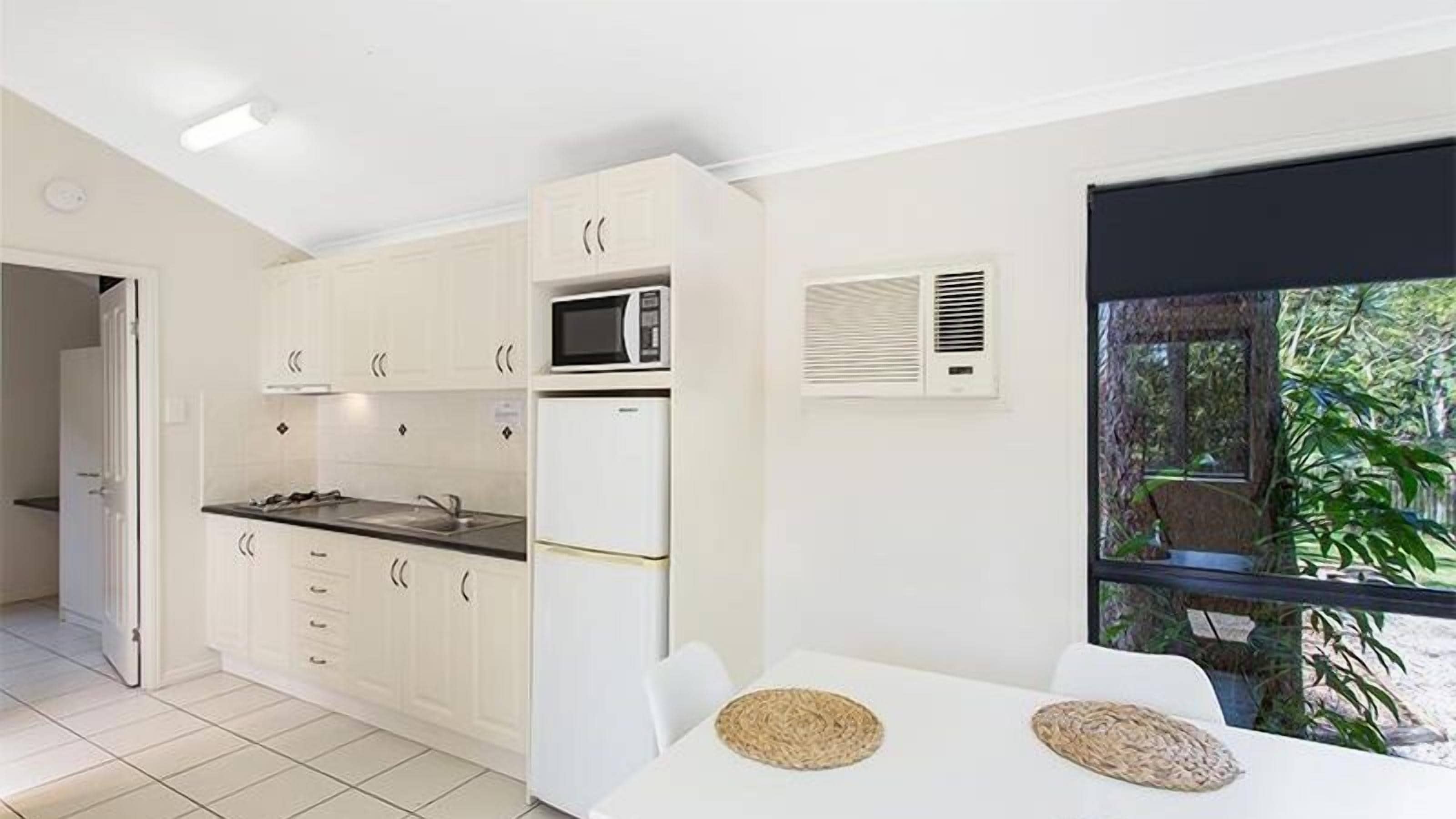 Eine moderne Küche mit weißen Schränken, einer Mikrowelle und einem Kühlschrank. Es gibt eine Essecke mit Tisch und Stühlen, und ein Fenster mit Blick auf Grünfläche.