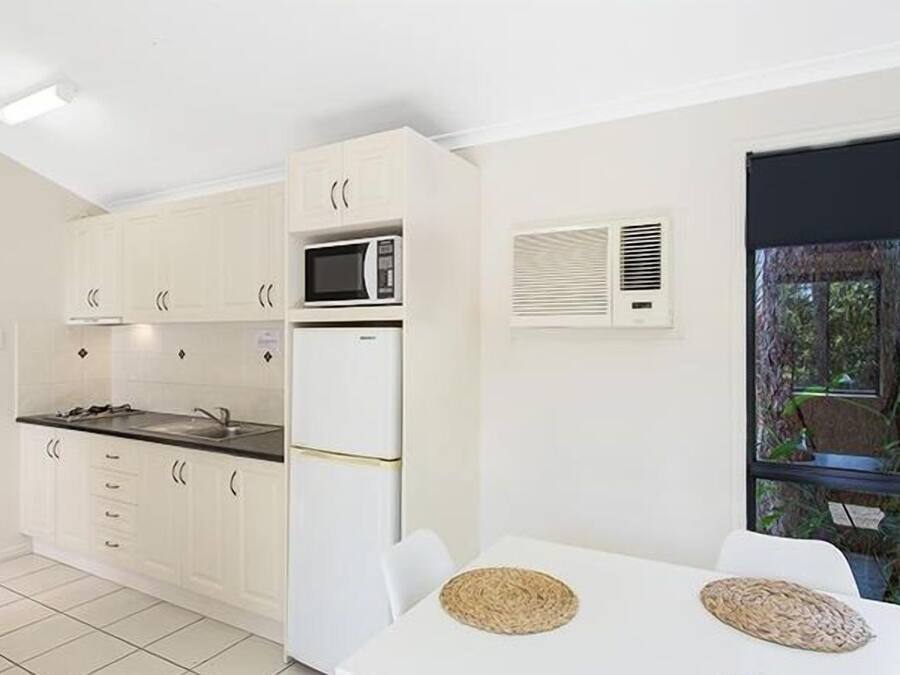 A modern kitchen with white cabinets, a microwave, and a refrigerator. There is a dining area with a table and chairs, and a window with a view of greenery outside.