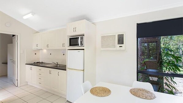 A modern kitchen with white cabinets, a microwave, and a refrigerator. There is a dining area with a table and chairs, and a window with a view of greenery outside. - Landsborough Holiday Park (Landsborough)