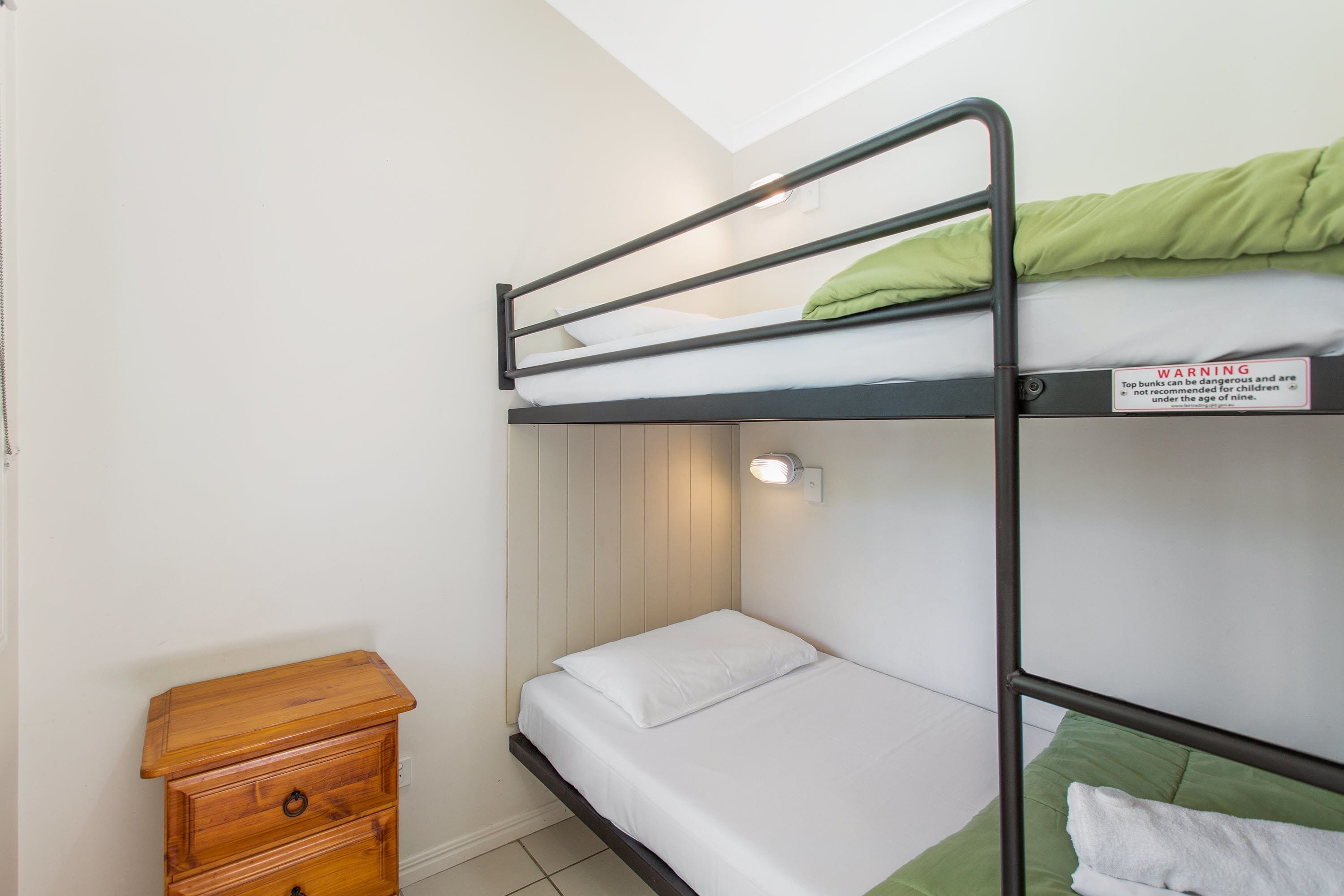 A bunk bed with a warning sign, a wooden nightstand, and a wall-mounted light fixture.