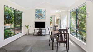 Un salon moderne doté d’un téléviseur à écran plat, d’une table à manger avec des chaises, de grandes fenêtres offrant une vue sur la verdure et d’un tableau accroché au mur.