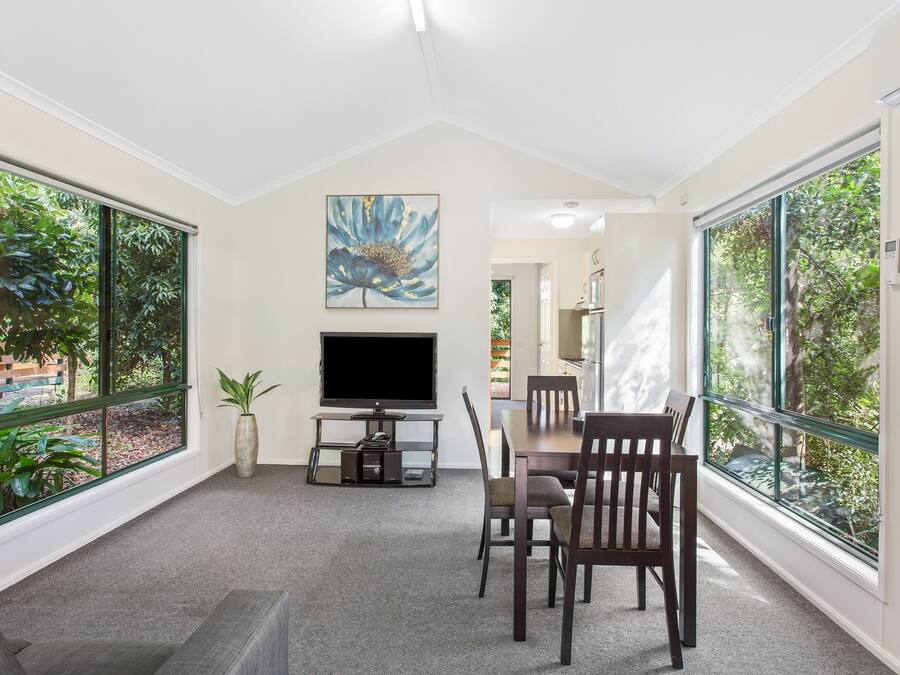 A modern living room with a flat-screen TV, a dining table with chairs, large windows with a view of greenery, and a painting on the wall.