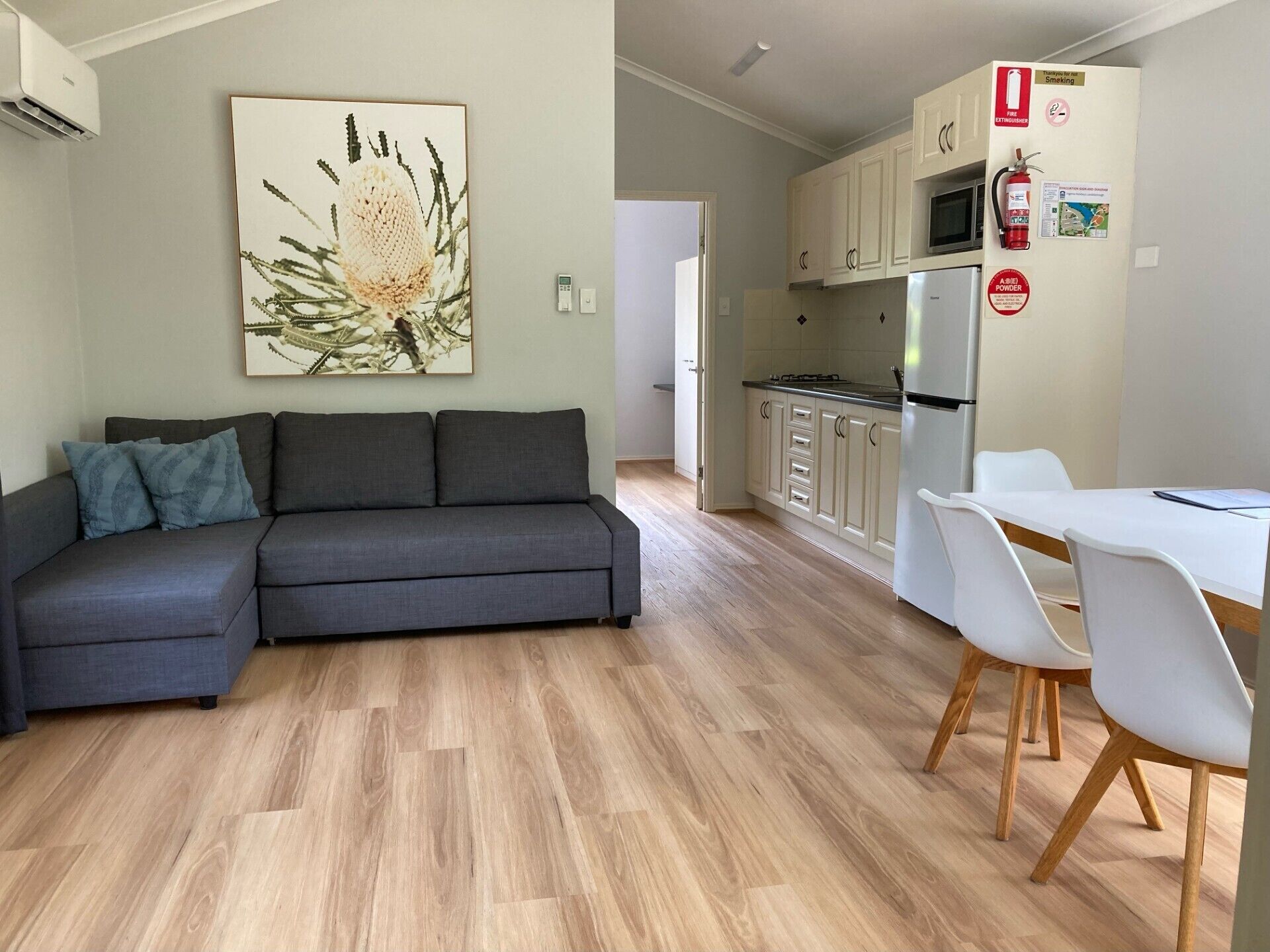 A modern living room with a grey sofa, a dining table with white chairs, and a kitchen area with white cabinets and a microwave.