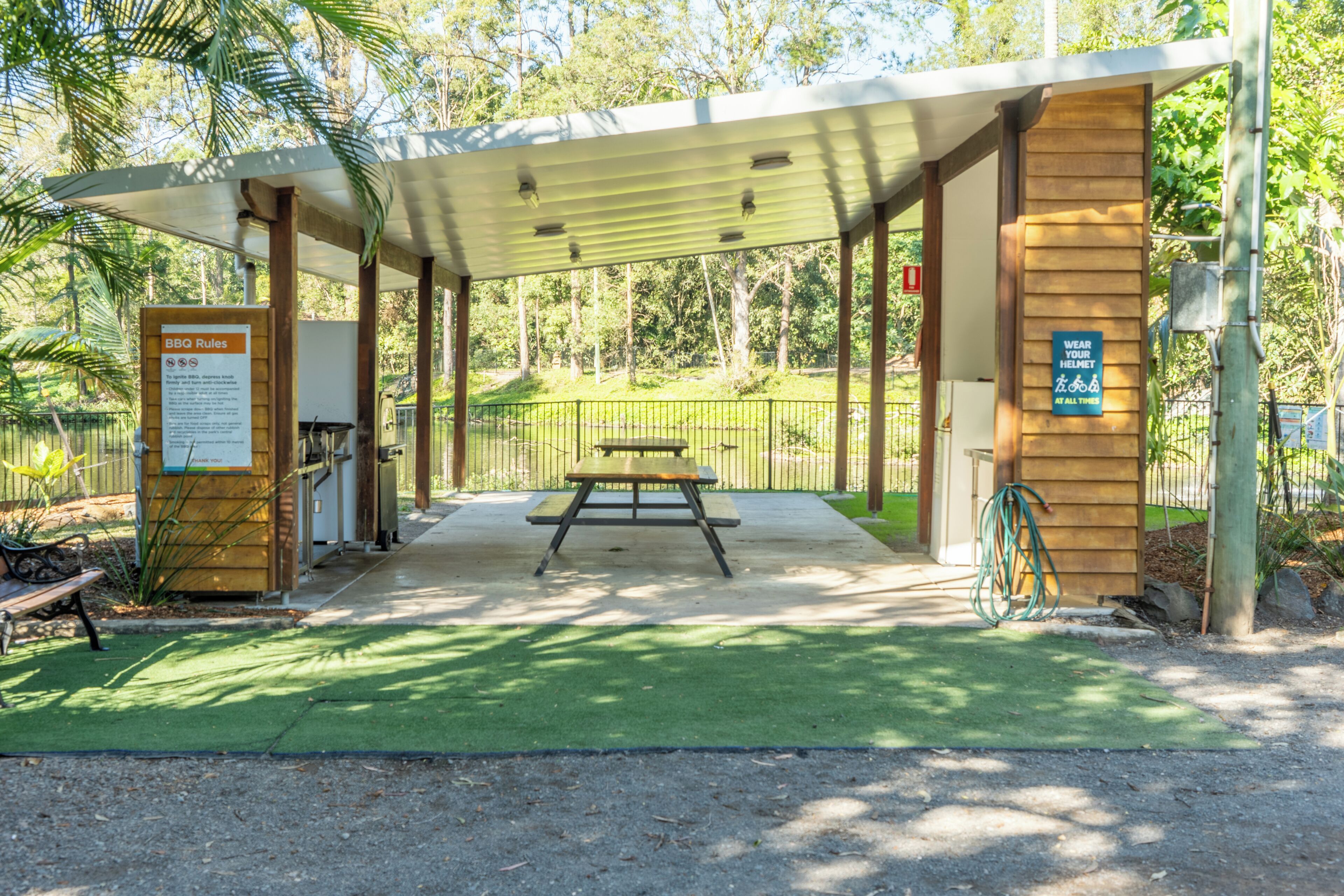 A covered picnic area with a table and benches, surrounded by a fence and greenery.