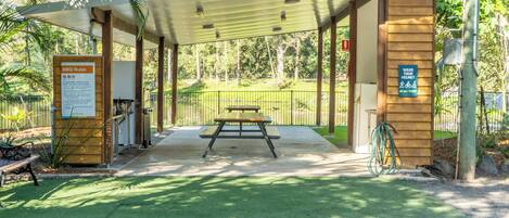 A covered picnic area with a table and benches, surrounded by a fence and greenery.